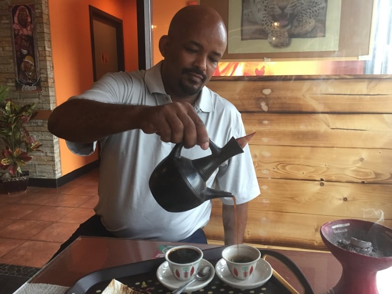 A Man Sits In A Booth In A Restaurant And Pours Coffee Into Cups On A Tray From An Ethnic Looking Pot.  A Container Containing What Looks Like Smoldering Ash Sits On The Table Next To Him.